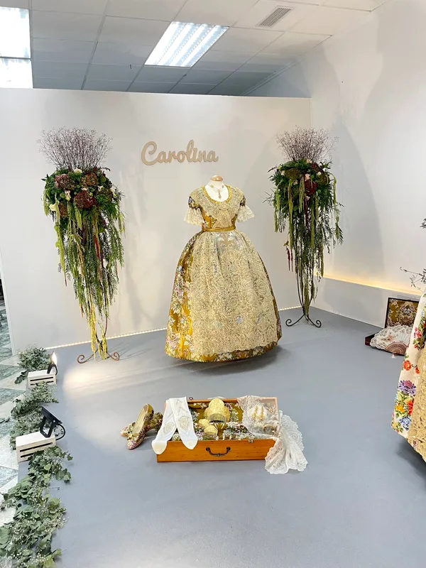 Traje de fallera de Carolina Martínez Serena expuesto entre dos centros de flores, con su nombre en la pared
