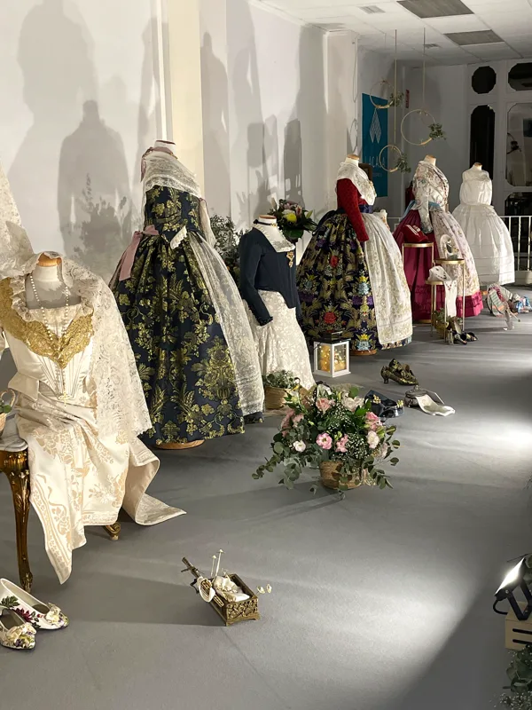 Exposición de trajes de fallera a lo largo de la sala de Lacasa, con flores y complementos en el suelo