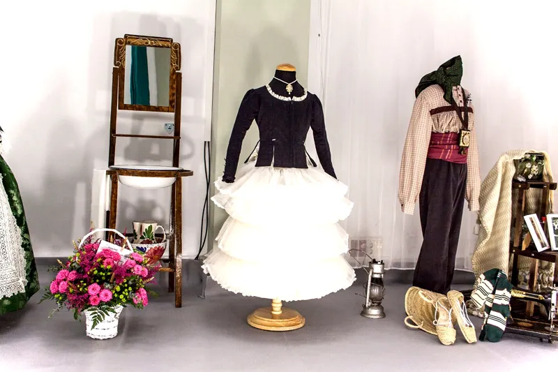 Trajes de valenciana y de saragüell expuestos con flores y una silla antigua