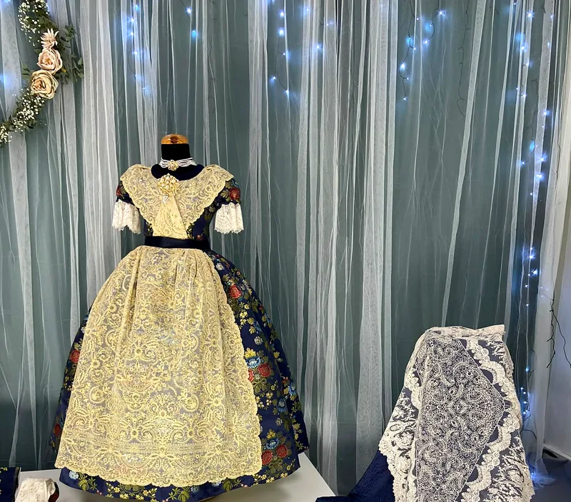 Traje de fallera infantil con delantal de encaje y mantilla, ante una cortina con luces