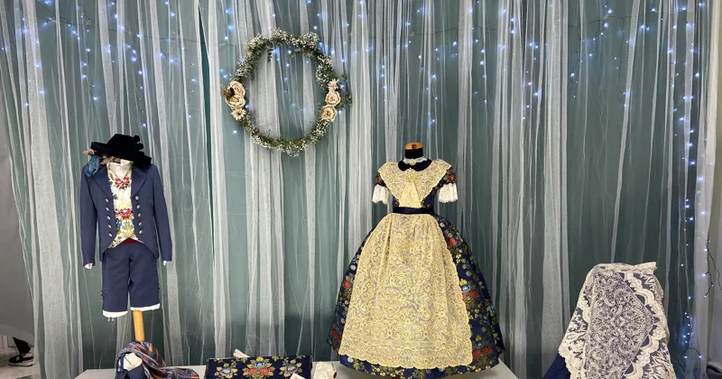Traje de saragüell y traje de fallera infantil expuestos ante una cortina con luces y una corona de flores