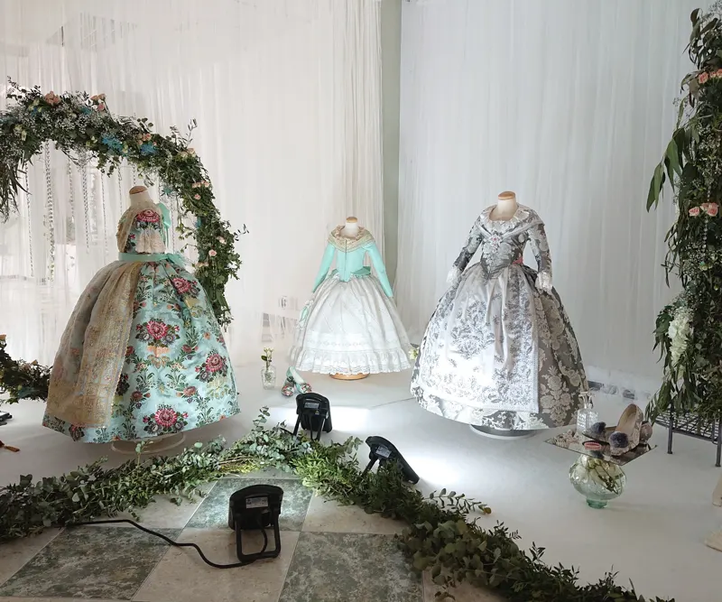 Trajes de fallera expuestos en maniquíes, con un arco de flores y focos en el suelo